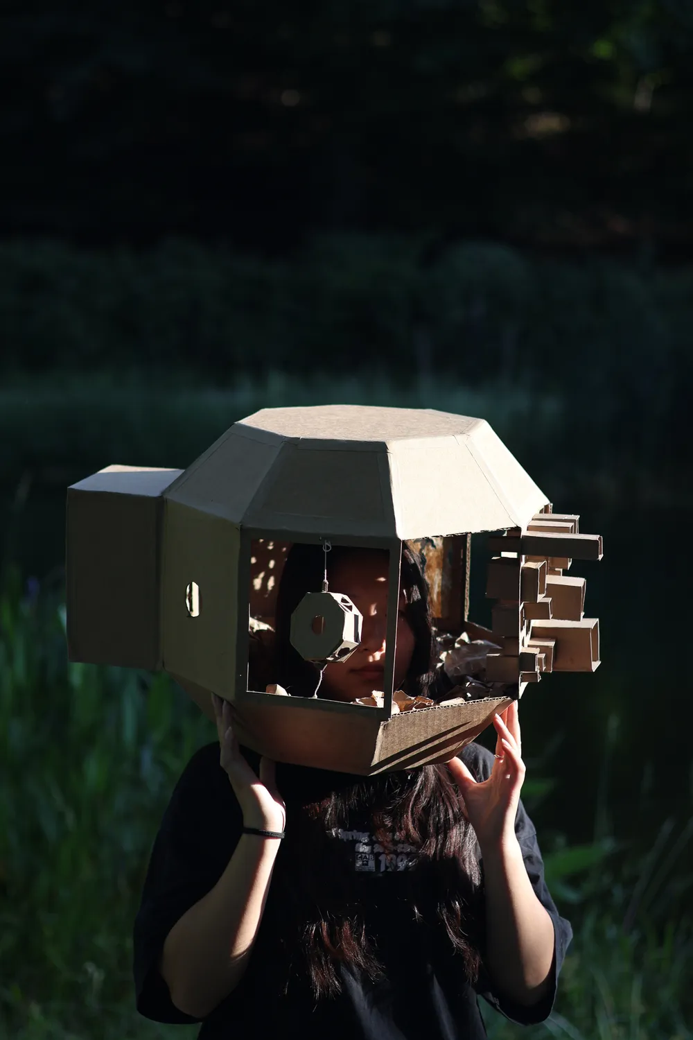 A faceted cardboard headpiece worn over someone's head in a dark field: a box lens juts from one side, a hanging octagonal disc sits in the open front beside their face, and stepped blocks project from the other side.